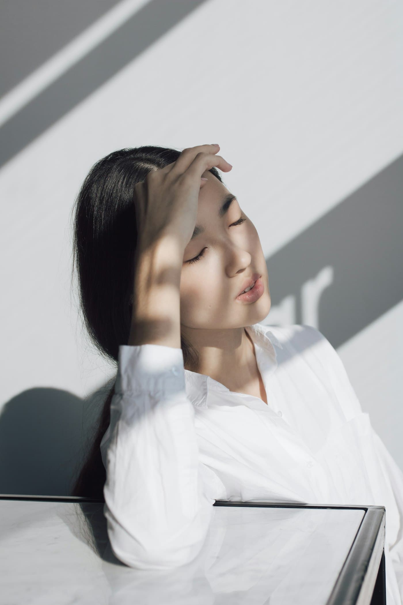 Mulher de camisa branca com a mão nos cabelos, iluminada por luz natural e sombras suaves