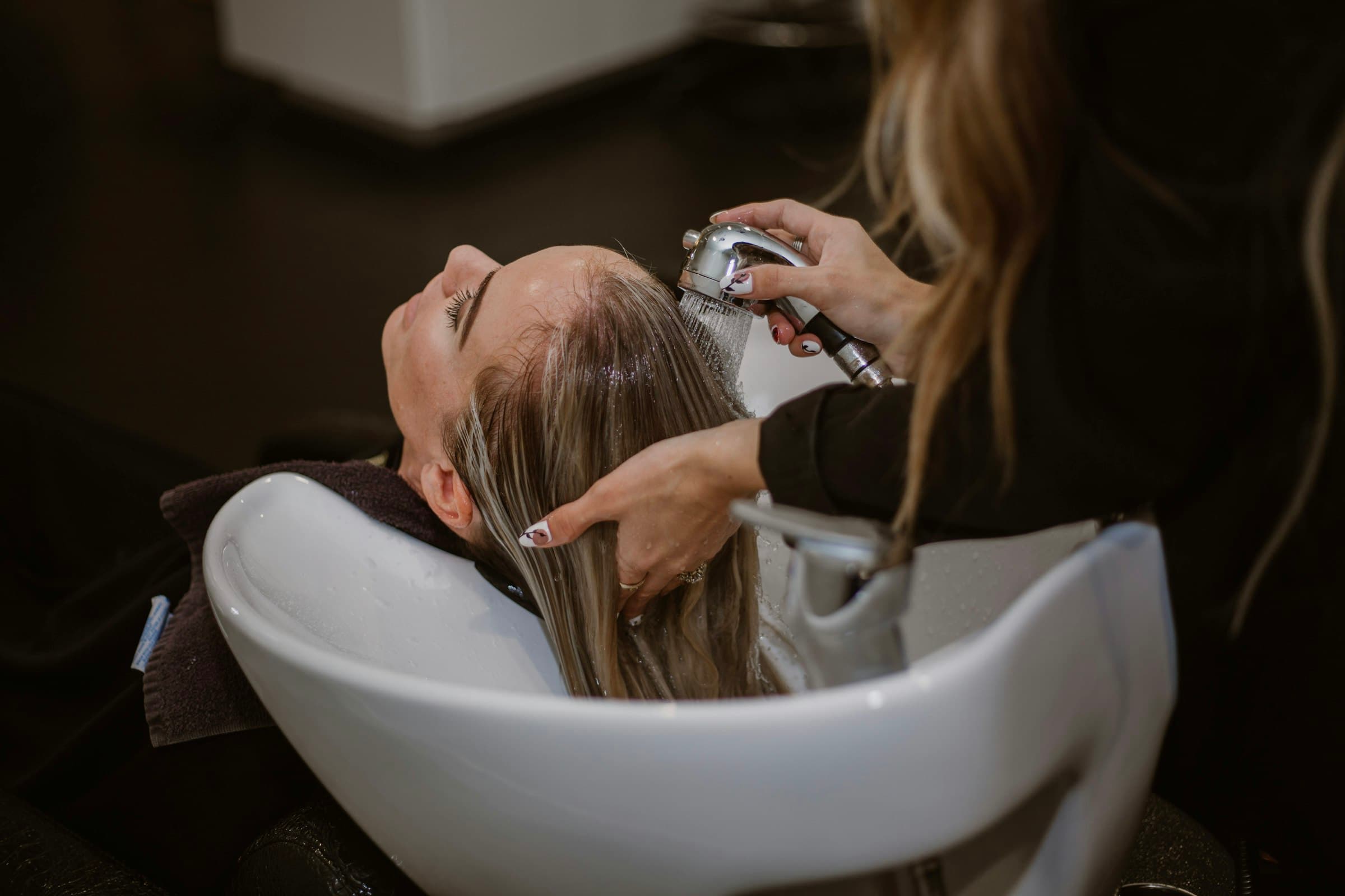 Cliente de olhos fechados recebendo lavagem de cabelo no lavatório, em luz baixa e acolhedora